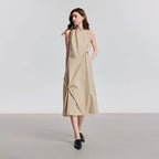 Model draagt een beige mouwloze midi jurk met ronde hals en gedrapeerde details.