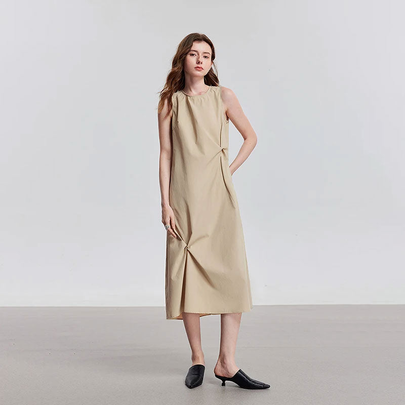 Model draagt een beige mouwloze midi-jurk met ronde hals en plooien aan de zijkant.