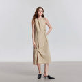 Model draagt een beige mouwloze midi-jurk met ronde hals en plooien aan de zijkant.