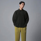 Man draagt een donkergrijs geplooid poloshirt met lange mouwen en losse pasvorm, gecombineerd met gele geplooide broek.