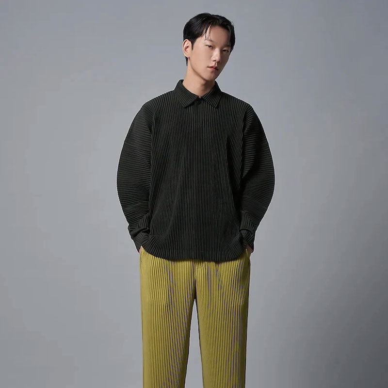 Man draagt een donkergrijs geplooid poloshirt met lange mouwen en losse pasvorm, gecombineerd met gele geplooide broek.