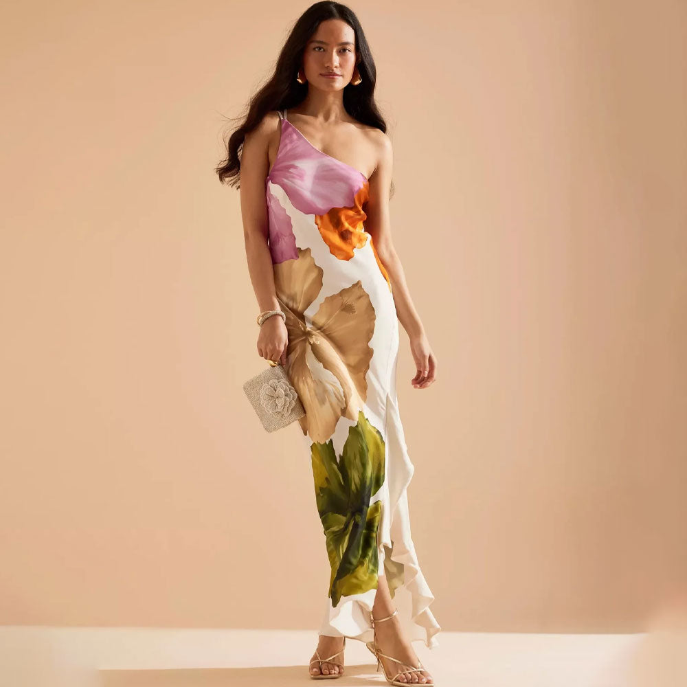 Elegante one shoulder maxi jurk met artistieke bloemenprint, golvende zoom en split, gecombineerd met hakken en tasje.